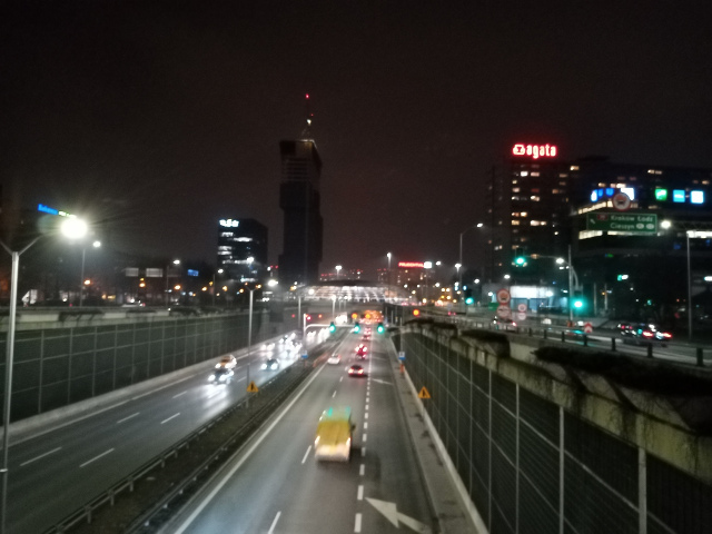 Zdjęcie wykonane w nocy, z punktu kilka metrów nad ruchliwą dwupasmową jezdnią. Widać kilka jadących po niej pojazdów. Po prawej stronie na budynku jest neonowy napis 'Agata'. W tle wysoki budynek o nieregularnym kształcie.