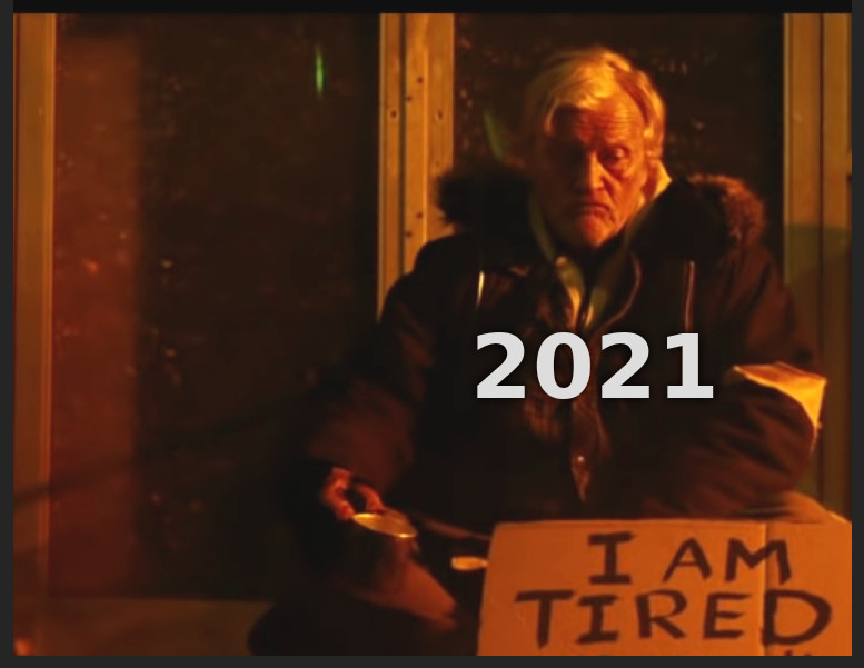 Stary bezdomny z filmu Hobo With a Shotgun, na którego nałożono napis 2021. Siedzi i trzyma kawałek kartonu, na którym napisano po angielsku 'Jestem zmęczony'.