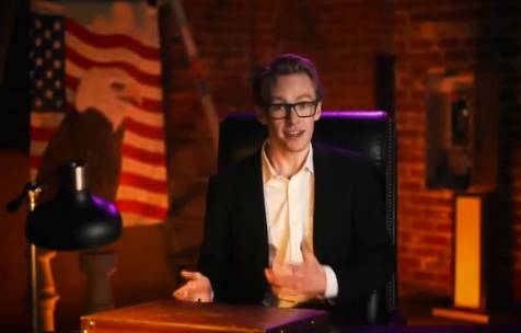 Kadr z filmu użytkownika Business Casual. Pokazuje blondyna w garniturze siedzącego za biurkiem i gestykulującego. Za jego plecami widać ceglane ściany, a po lewej powiewa amerykańska flaga z orłem.